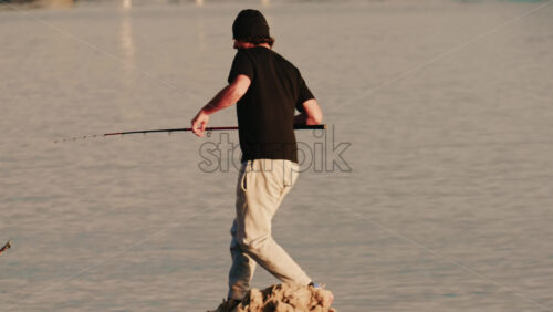 Video - Antibes, France - December 22, 2025: Rear view of a man fishing from the shoreline using a rod during warm evening light