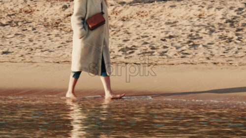 Video - Lower body view of a woman walking barefoot along the water's edge on a sandy beach
