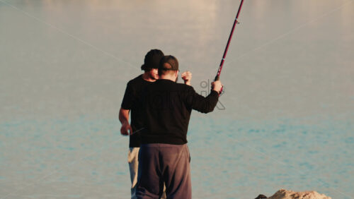 Video - Antibes, France - December 22, 2025: Two men standing on a rocky shore as one lifts a freshly caught fish from a fishing line