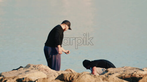 Video - Antibes, France - December 22, 2025: Two men crouching on coastal rocks while handling fishing equipment and a caught fish