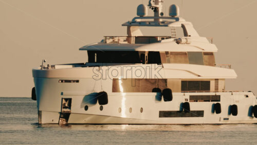 Video - Side view of a modern luxury yacht moving across calm sea water with a lighthouse in the distance