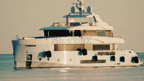 Video - Side view of a modern luxury yacht moving across calm sea water with a lighthouse in the distance