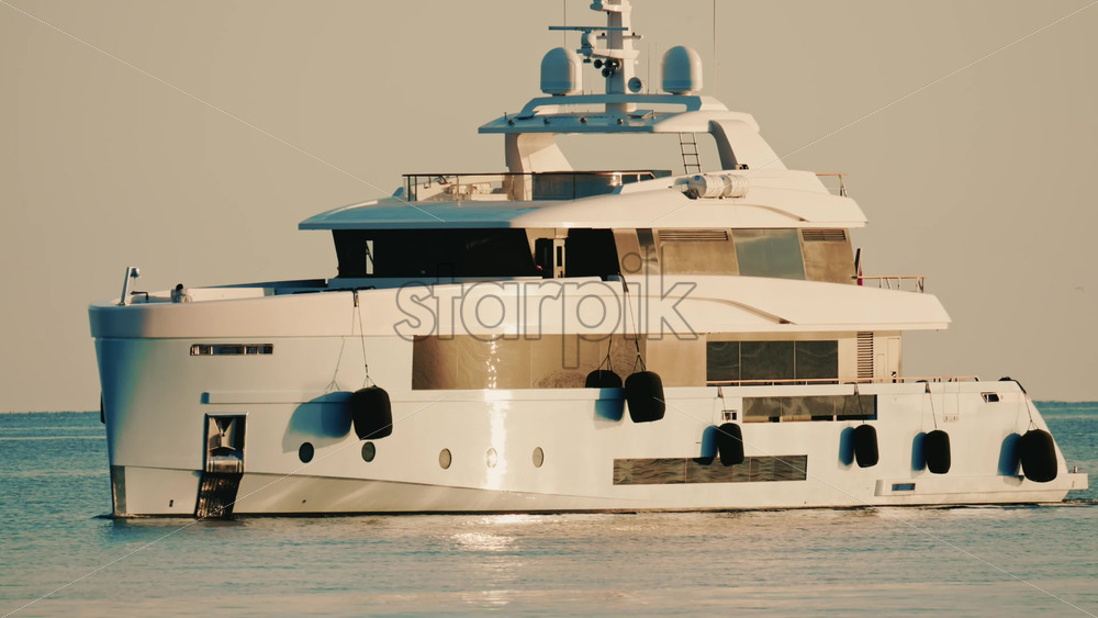 Video - Side view of a modern luxury yacht moving across calm sea water with a lighthouse in the distance