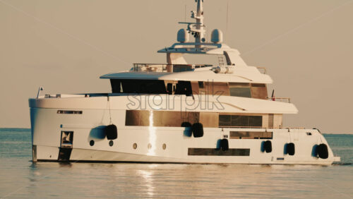 Video - Side view of a modern luxury yacht moving across calm sea water with a lighthouse in the distance