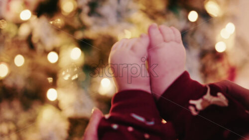 Video - Close up of baby feet wearing festive pajamas held in front of a softly lit Christmas tree