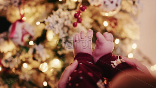 Video - Close up of baby feet wearing festive pajamas held in front of a softly lit Christmas tree