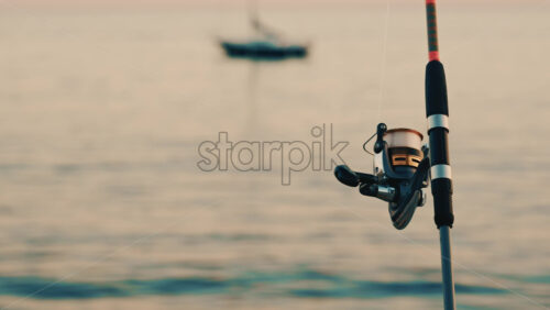 Video - Close up of a fishing rod and reel positioned near the sea, with a blurred boat in the background