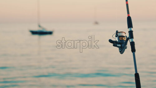 Video - Close up of a fishing rod and reel positioned near the sea, with a blurred boat in the background
