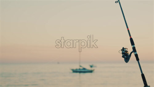 Video - Close up of a fishing rod and reel positioned near the sea, with a blurred boat in the background
