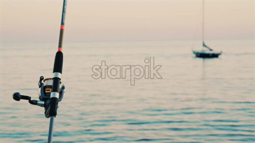 Video - Close up of a fishing rod and reel positioned near the sea, with a blurred boat in the background