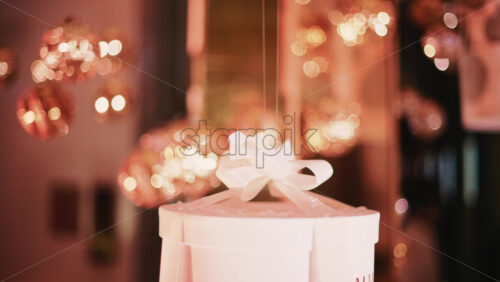 Video - Detail shot of a round white gift box topped with a satin ribbon, surrounded by softly blurred festive decor and warm lights