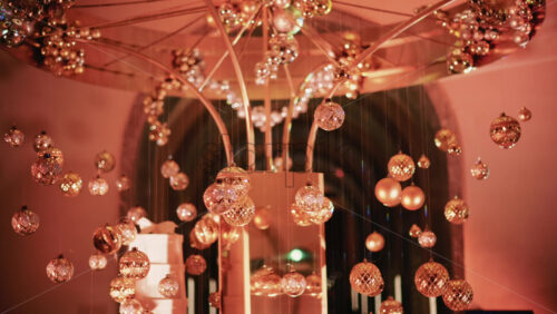 Video - Golden Christmas baubles hanging from a structure alongside a suspended white gift box, with people blurred in the background