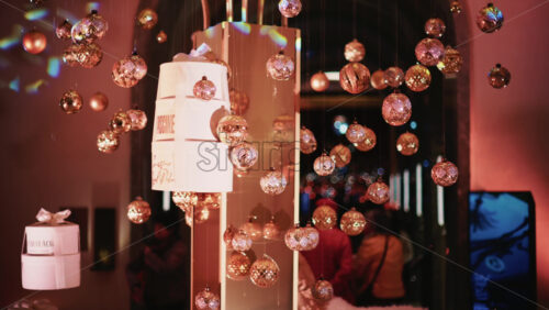 Video - Golden Christmas baubles hanging from a structure alongside a suspended white gift box, with people blurred in the background