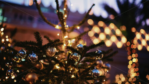 Video - A Christmas tree decorated with metallic ornaments and fairy lights, set outdoors with blurred lights and greenery behind it