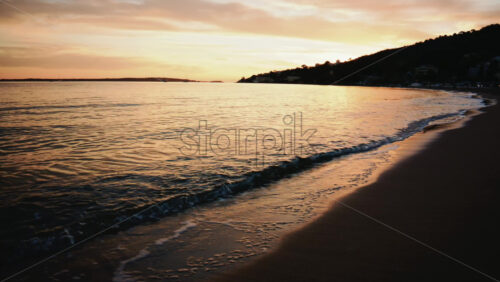Video - Gentle sea waves rolling onto a sandy beach during sunset, with anchored sailboats visible on the horizon