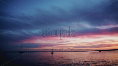 Video - Wide shot of calm sea with sailboats anchored offshore beneath a colorful sunset sky with pink and blue clouds