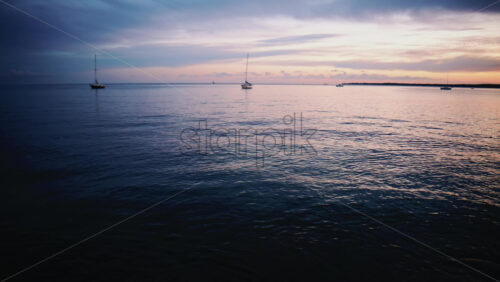 Video - Wide shot of calm sea with sailboats anchored offshore beneath a colorful sunset sky with pink and blue clouds