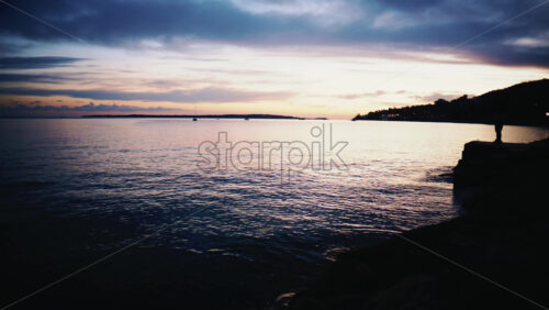 Video - Wide shot of calm sea with sailboats anchored offshore beneath a colorful sunset sky with pink and blue clouds