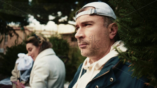 Video - Close up of a man wearing a jacket and hoodie, standing thoughtfully among evergreen Christmas trees in an outdoor market setting