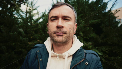 Video - Portrait shot of a man looking away from the camera, framed by dense green fir trees, with soft natural daylight and shallow depth of field