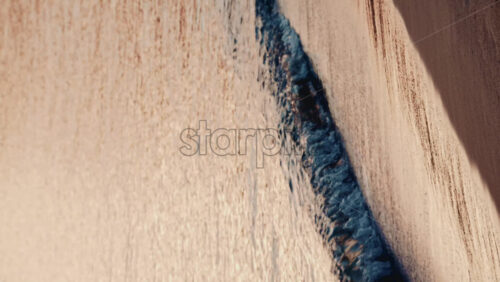 Video - Gentle sea waves rolling onto a sandy beach during sunset, with anchored sailboats visible on the horizon. Vertical