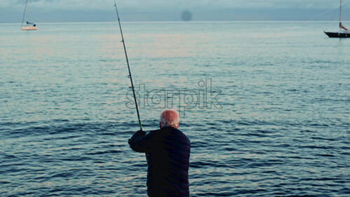 Video - Cannes, France - December 23, 2025: Adult man standing on a rocky shoreline while fishing, holding a long rod directed toward calm coastal waters