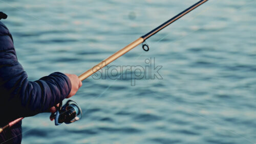 Video - Adult man standing on a rocky shoreline while fishing, holding a long rod directed toward calm coastal waters