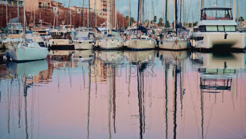 Video - Cannes, France - December 23, 2025: Wide shot of a marina filled with moored boats, their masts reflected in soft pink and blue water at sunset