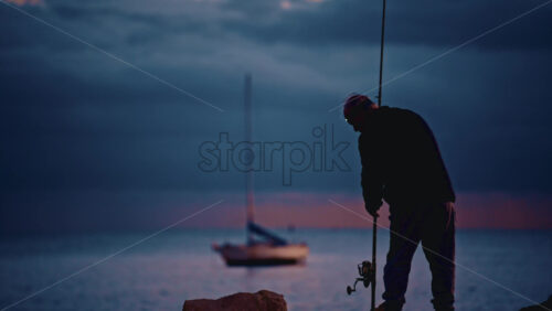 Video - Silhouette of an elderly man fishing from the shore at dusk, with a sailboat anchored in the distance