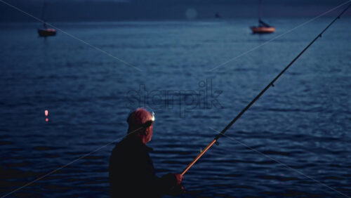 Video - Cannes, France - December 23, 2025: Side view of a fisherman wearing a head light casting his line into calm sea waters during blue hour