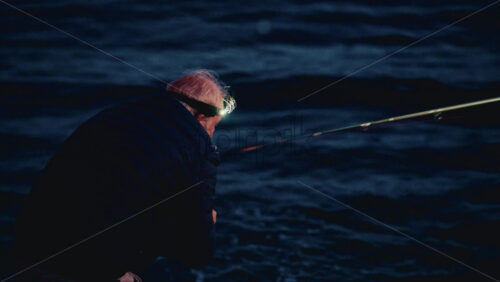 Video - Cannes, France - December 23, 2025: Side view of a fisherman wearing a head light casting his line into calm sea waters during blue hour