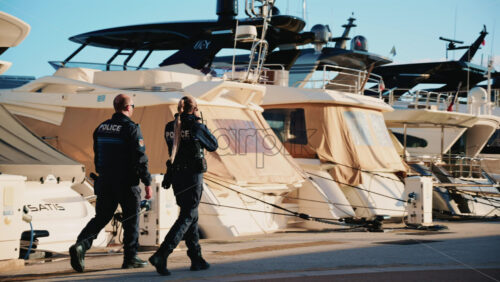 Video - Cannes, France - December 23, 2025: Two uniformed police officers walking along a marina promenade with luxury yachts docked in the background