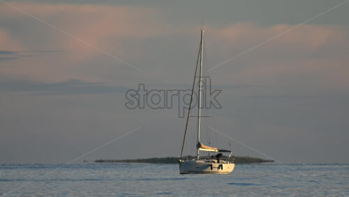 Video - A lone sailboat floating peacefully on calm sea waters, with a soft pastel sky and distant coastline on the horizon