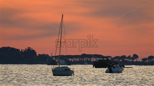 Video - Small boats anchored offshore during sunset, with warm orange and pink tones reflecting on the sea and silhouettes of trees in the distance