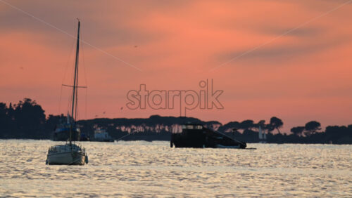 Video - Small boats anchored offshore during sunset, with warm orange and pink tones reflecting on the sea and silhouettes of trees in the distance