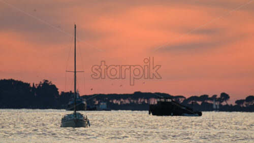 Video - Small boats anchored offshore during sunset, with warm orange and pink tones reflecting on the sea and silhouettes of trees in the distance