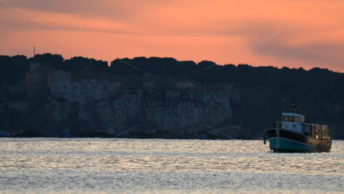 Video - Traditional fishing boat drifting on gentle waves near a rocky coastline, under a dramatic dusk sky with warm colors
