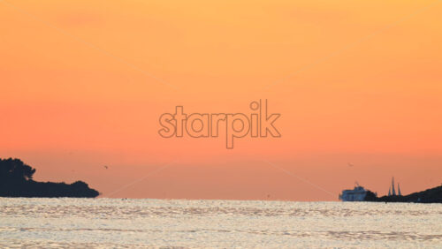 Video - Wide seascape showing a distant vessel on the horizon beneath an orange sunset sky, with calm water
