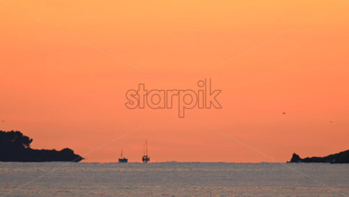 Video - Wide shot of calm sea under a colorful sunset sky, with distant islands and boats silhouetted on the horizon