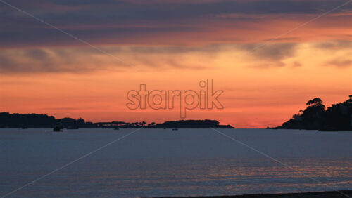Video - Wide shot of calm sea under a colorful sunset sky, with distant islands and boats silhouetted on the horizon