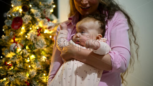 Video - Mother holding a baby indoors next to a decorated and illuminated Christmas tree