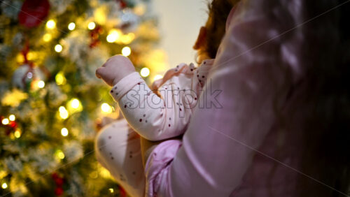 Video - Mother holding a baby indoors next to a decorated and illuminated Christmas tree