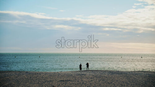 Video - Two people standing near the shoreline, looking out at the sea under a soft evening sky