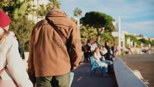 Video - Cannes, France - December 23, 2025: Pedestrians walking along La Croisette on a sunny day, with palm trees, benches, and the Mediterranean Sea in the background