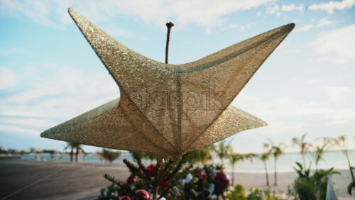 Video - Close up of a golden Christmas star decoration with blurred palm trees and beach in the background