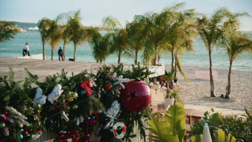 Video - Christmas garlands and ornaments decorating a seaside structure, with palm trees and sandy beach visible behind