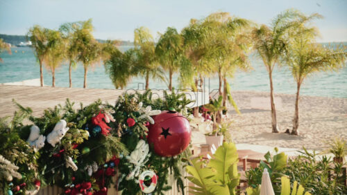 Video - Christmas garlands and ornaments decorating a seaside structure, with palm trees and sandy beach visible behind