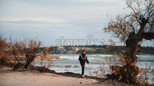 Video - Cannes, France - December 23, 2025: Man walking along a sandy beach framed by coastal vegetation, with Ile Sainte Marguerite visible across the water