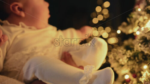 Video - A close, warmly lit shot of a baby dressed in a soft white outfit, being gently held by a mother's hands