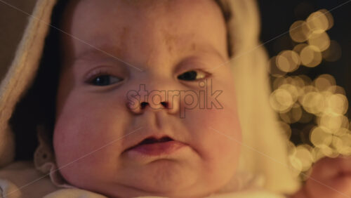 Video - Close up of a baby's face with blue eyes and smooth, rosy skin with festive golden bokeh lights in the background
